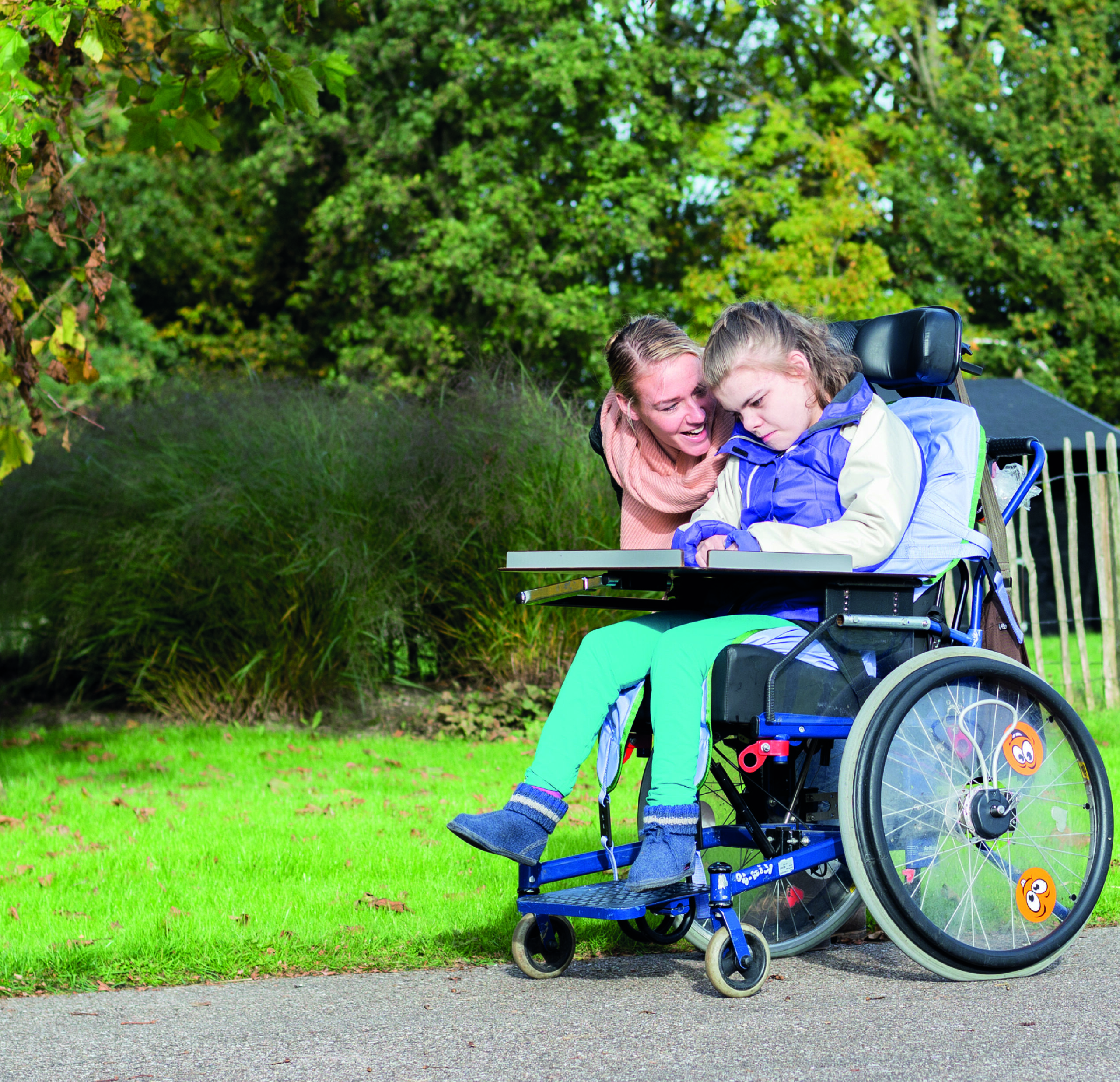 A mother kneeling beside her daughter, who uses a wheelchair, in a park